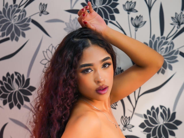 MadisonDawson has curly dark red hair, tan skin, and red lipstick. She's looking at the camera with her arm raised, showing off her bare shoulders and a nose ring.
