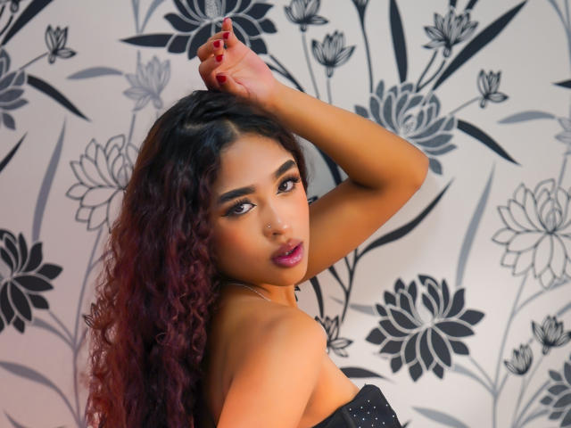 MadisonDawson with curly, purple-tinted hair, raised arm, dark lipstick, and red nails, wearing a black strapless top, looking over her shoulder with seductive eyes.
