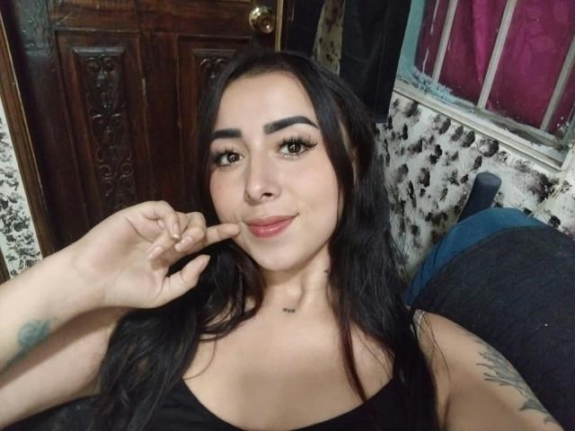 AnneHoneyy with long black hair, thick eyebrows, and full lips, pointing at her chin with a slight smile, wearing a black top, showing tattoos on her arm and chest.