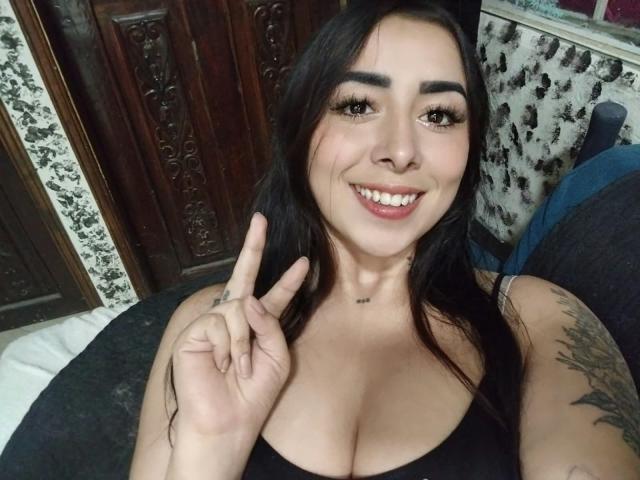 AnneHoneyy with dark hair, big tits, and a smile, making a peace sign with her right hand, showing tattoos on her left arm, in a selfie.
