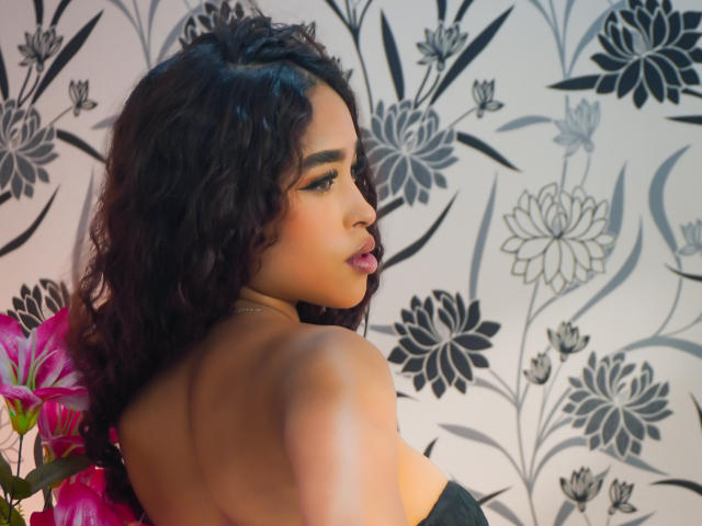 MadisonDawson with curly black hair, dark skin, and pink lipstick, looking over shoulder, showing bare back and black strap, next to pink flowers, floral wallpaper in background.