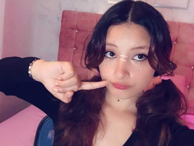 ValentinaRoseX with pouty lips, dark wavy hair in pigtails, black top, white face markings, pointing finger at nose, pink tufted headboard, and a bracelet on her wrist.