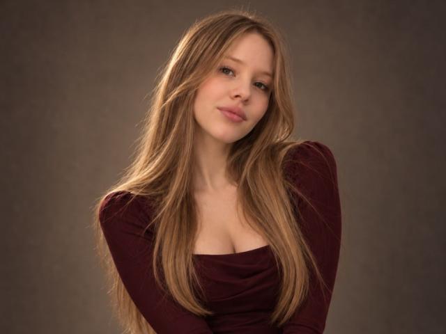 LanieDistel has long, straight, blonde hair, wearing a maroon top with a low-cut neckline that shows her small, perky tits. She's looking slightly to the side with a subtle smile.