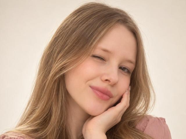 LanieDistel has wavy blonde hair, fair skin, and is winking with a cute smile. She's touching her cheek with her hand, wearing a pink top.