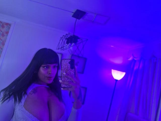 TeylorJenkins with long black hair, bangs, and a white lace top, holding a phone with a pink glitter case, showing her big tits in a purple-lit room.
