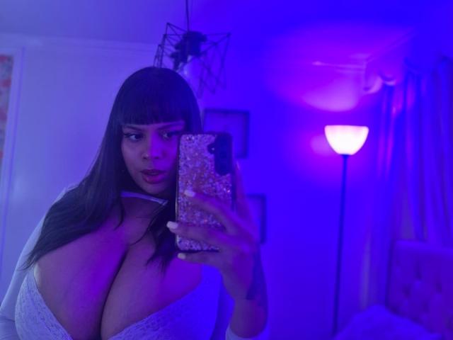 TeylorJenkins with big boobs, wearing a white lace top, taking a mirror selfie in a blue-lit room, holding a glittery phone, looking at the camera with a slight pout.