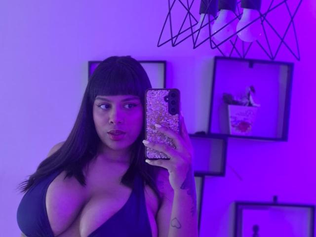 TeylorJenkins with large tits, holding phone with glittery case, wearing black top, purple lighting, straight black hair with bangs, looking at mirror, purple background.
