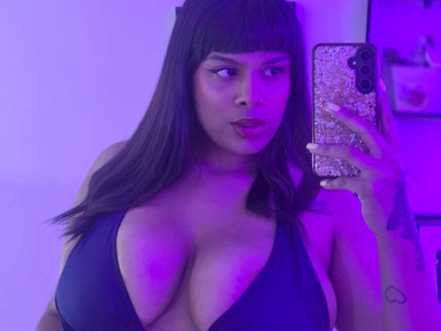 TeylorJenkins with big tits, wearing a black halter top, taking a mirror selfie with purple lighting, holding a glittery phone, looking to the side with dark lipstick.