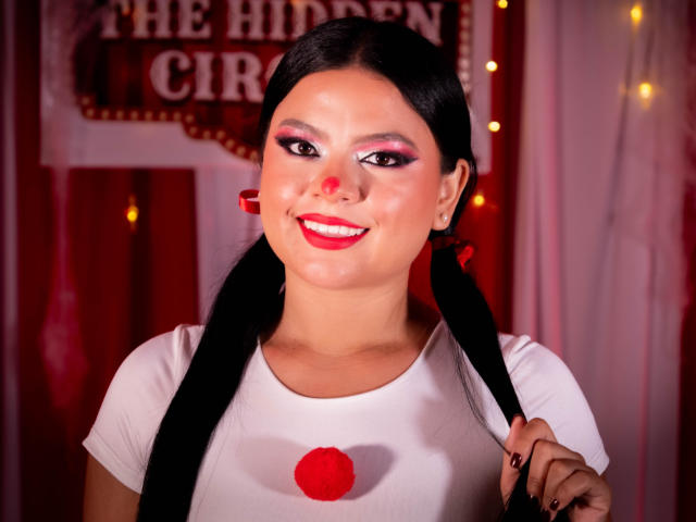 MelanyPorto with black pigtails, white shirt, red nose, pink eyeshadow, red lipstick, holding a red pom-pom, smiling, red background, fairy lights, slight blush, medium tits.