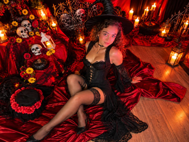 Arianna Blake, déguisée en sorcière avec un costume noir, un corset à lacets, une jarretière et des bas, assise sur du satin rouge, portant un chapeau de sorcière, avec des bougies, des crânes et des fleurs, montrant ses seins..