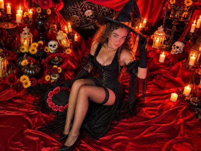 Arianna Blake, avec des cheveux bouclés, portant un chapeau de sorcière, une robe noire, des bas résille et des gants, assise sur du satin rouge, entourée de bougies, de crânes et de fleurs, et ayant une allure séduisante..