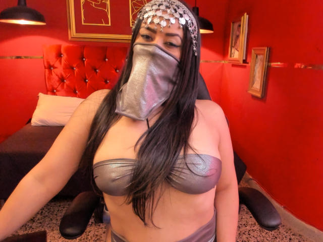 Anissa Ali mit langen, schwarzen Haaren, die einen silbernen Kopfschmuck, eine Metallmaske und ein metallenes BH-Top trägt, wobei ihre großen Brüste und ihr kurvige Körper sichtbar sind, in einem rot gestrichenen Raum..