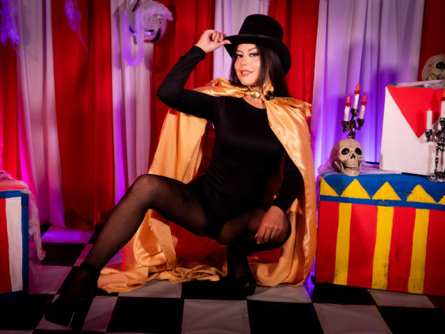 ElectraRoss está en cuclillas, vestida con lencería negra, medias transparentes y un sombrero negro, con una capa dorada, mostrando su trasero, en un escenario temático de Halloween con un cráneo y velas..