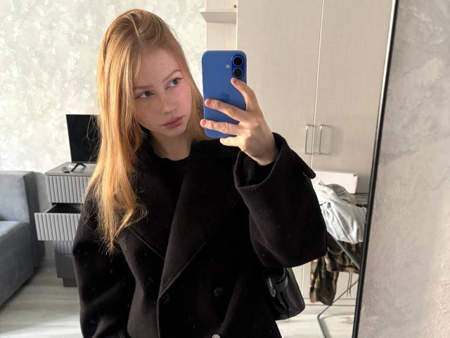 CorlissCrunkleton with blonde hair, fair skin, wearing a black coat, holding a blue phone, taking a mirror selfie, showing a slight pout, in a modern room.