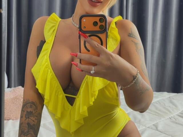 NicolSNaya trägt ein gelbes, gerafftes Einteiler-Kostüm und zeigt auf einem Spiegel-Selfie große Brüste, mit roten Nägeln, einem Ring und Tattoos. Sie hält ein orangefarbenes Telefon, ihre Lippen sind leicht geöffnet..