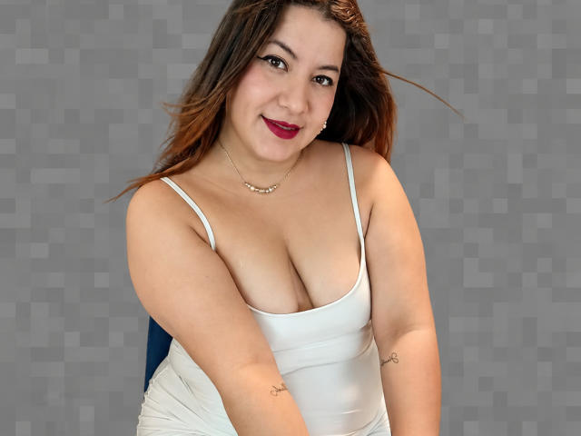 SalomeVos, avec de grandes seins, portant un débardeur blanc, souriant avec du rouge à lèvres, des cheveux bruns et des tatouages sur les bras, sur un fond gris. Elle porte également un collier..
