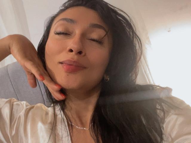 AmeliRoux with her eyes closed, dark wavy hair, pink lipstick, and a slight smile, wearing a white shirt, hand on cheek, red nails, and a silver necklace.