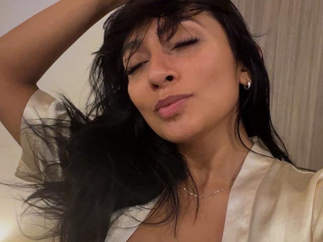 AmeliRoux, avec ses cheveux noirs, les yeux fermés, les lèvres pincées, portant une robe blanche en soie, une main dans les cheveux, de petites boucles d'oreilles, un collier discret, une peau douce, une expression détendue..