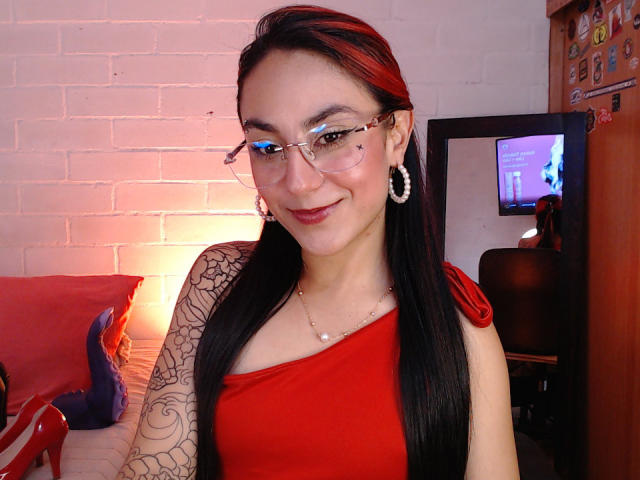 AmyStreetwise69 trägt ein rotes Kleid, eine Brille, Reifenohrringe, einen schwarzen Tattoostreifen am Arm und einen roten Stirnband. Sie lächelt in die Kamera und zeigt in dem Foto ihre Brüste und ihren Hintern..