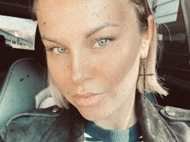 BelleLisaG con un corte de pelo rubio, ojos verdes, maquillaje sutil, vistiendo una chaqueta de cuero negra y un arete de cruz, con una expresión ligeramente sensual y seductora..