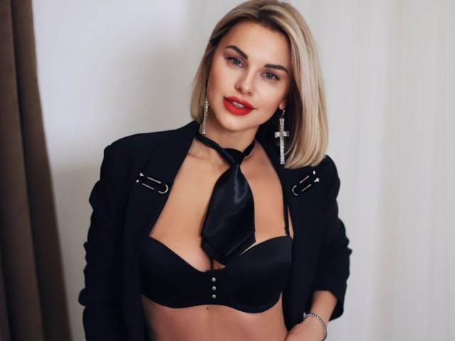 BelleLisaG con caschetto biondo, rossetto rosso, reggiseno nero e blazer con cravatta, che mostra il seno, con orecchini a croce, dall'aspetto sexy..