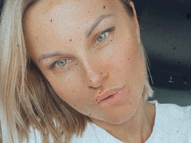 BelleLisaG hat blondes Haar, blaue Augen und macht ein schmollendes Gesicht mit einem kleinen Schnitt auf ihrer Unterlippe. Sie trägt ein weißes Hemd und hat kleine, dunkle Muttermale..