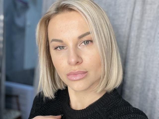 BelleLisaG with blonde bob, light skin, subtle makeup, pink lips, and black knit sweater, looking directly at camera, holding sweater collar, green eyes, slight smile.