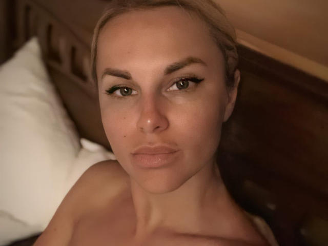 BelleLisaG mit vollen Lippen, grünen Augen und blonden Haaren, die nach oben blickt, oben ohne, wobei ihre nackten Schultern zu sehen sind, mit einem hölzernen Kopfteil im Hintergrund. Ihr Gesicht ist mittig platziert..