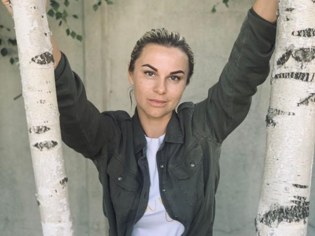 BelleLisaG with arms raised, holding white birch trees, wearing a green jacket over a white tee, brown hair in a messy bun, green eyes, and a slight smile.