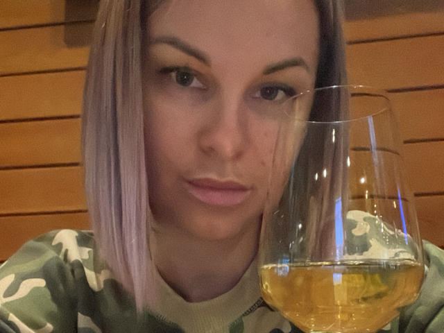BelleLisaG mit einem rosa-braunen Bob, hält ein Glas Weißwein, leicht gerötete Lippen, trägt ein Tarnhemd, Hintergrund mit Holzpaneelen. Ihre Brüste sind klein, sie schaut in die Kamera..