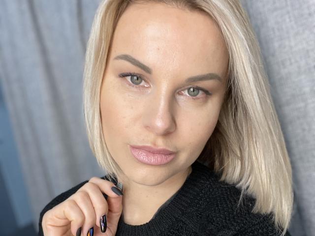 BelleLisaG hat blondes Haar, grüne Augen und rosafarbene Lippen. Sie trägt einen schwarzen Pullover und hat schwarze Fingernägel. Sie schaut direkt in die Kamera und hat leicht geschlossene Lippen..
