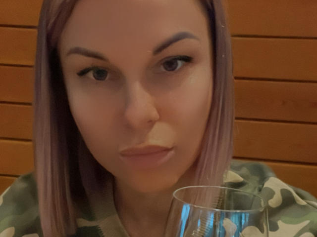 BelleLisaG with purple hair, raised eyebrows, and slightly pouty lips, holding a glass, wearing a green and white camouflage shirt. Her face is centered in the shot.