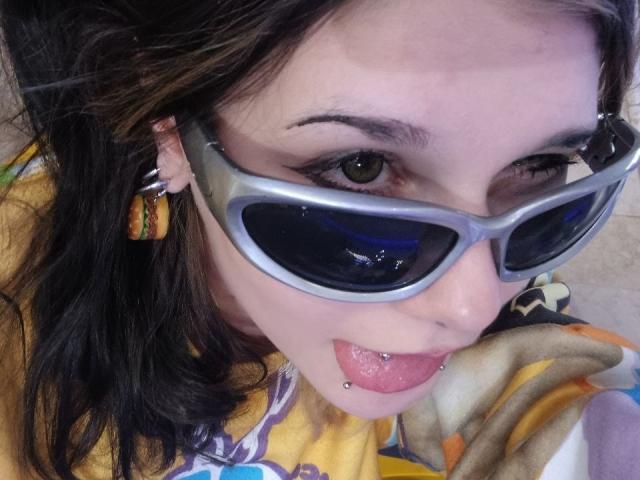 ShenaBatrum, com óculos de sol cinzas, camisa amarela e cabelo castanho, com a língua de fora e um piercing no lábio, usando brincos coloridos e olhando para a câmera..