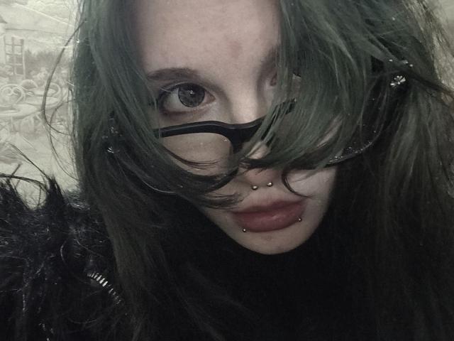 ShenaBatrum ha i capelli verdi, occhiali neri e piercing sul naso e sul labbro inferiore, con rossetto scuro, e guarda la fotocamera con un'acconciatura leggermente disordinata..