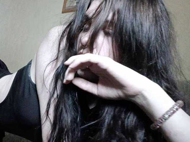 ShenaBatrum mit langem, zerzaustem, schwarzen Haar, das ihr Gesicht bedeckt, trägt ein schwarzes Tanktop, ihre Hand bedeckt teilweise ihren Mund, sie hat ein Perlenarmband am Handgelenk, ihre Haut ist hell, sie trägt dezentes Make-up..