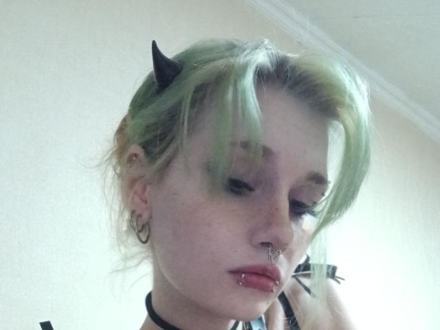 ShenaBatrum with green hair, black devil horns, nose ring, lip piercing, and hoop earrings, looking down with a black choker, wearing a black top, in a selfie.