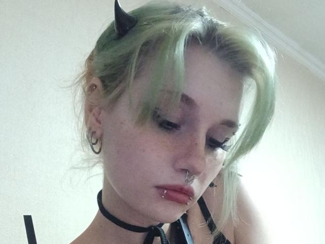ShenaBatrum with green hair, black horns, gold hoop earrings, black choker, nose ring, lip piercing, and looking down, showing freckles and eyelashes, wearing a black strap top.