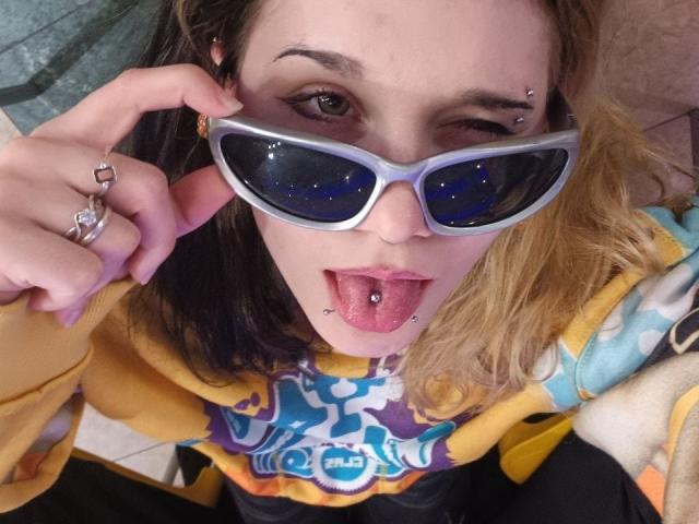 ShenaBatrum with a pierced tongue, blue-eyed, and nose, wearing reflective silver sunglasses, yellow and blue shirt, and multiple rings, sticking out her tongue and winking.