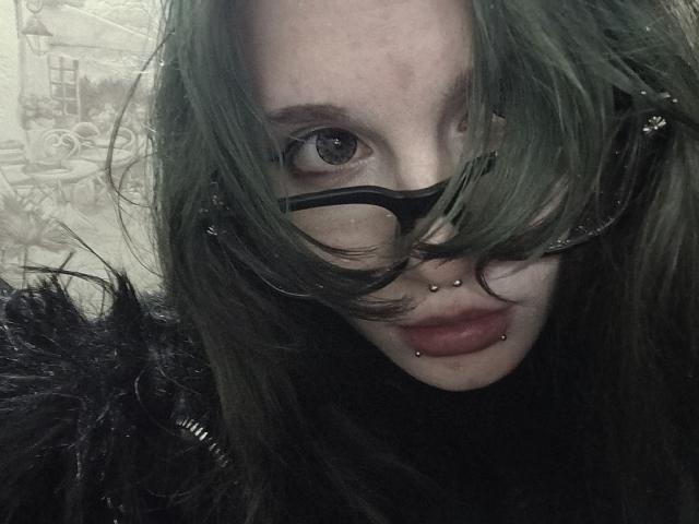 ShenaBatrum, avec des cheveux verts, des lunettes noires, un anneau au nez et un piercing aux lèvres, regardant la caméra, avec un col en fourrure noire, des yeux gris, des cheveux en désordre, un maquillage foncé, photo granuleuse..