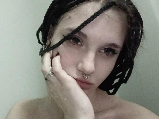 ShenaBatrum with wet black braids, nose piercings, and a ring, touching her cheek, looking at the camera, showing her pale skin and small tits.