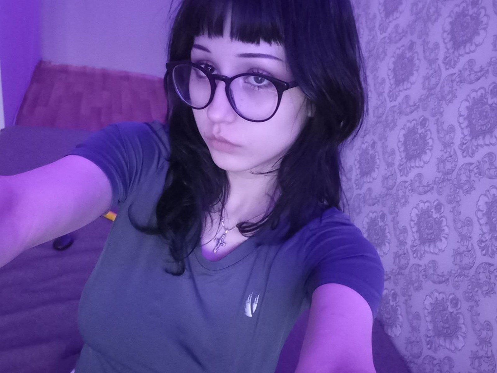 MarilynnGeales with black glasses, black wavy hair, and a blue-green shirt, showing her tits in a purple-lit selfie. She has a cross necklace and a serious expression.