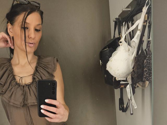 JoliDanielle with brown dress, holding phone, taking selfie in mirror. Her tits are small, brown hair in ponytail, glasses on head. White bra, black bag, clothes on right.