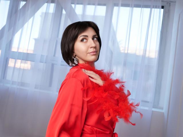 RoseWhisper with short black hair, wearing a red dress and red feather boa, looking to the side with a slight smile, earrings visible, in a well-lit room.
