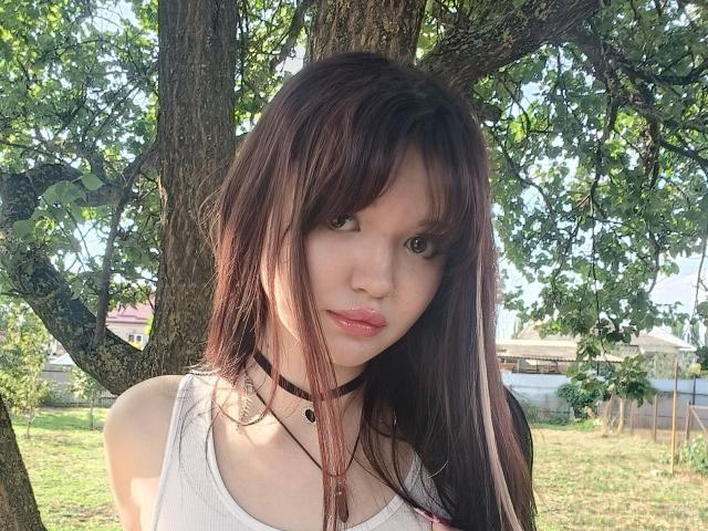 MistyEllstrom with long brown hair, small nose, pink lips, white tank top, black choker, and necklace, standing under a tree, looking directly at the camera.