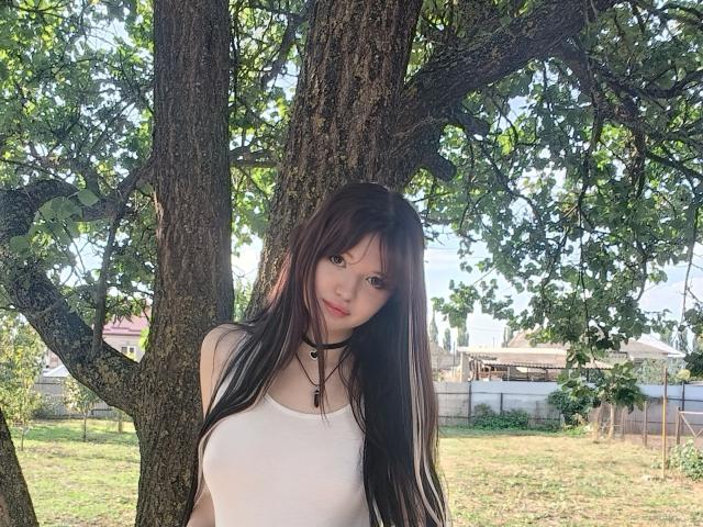 MistyEllstrom standing in front of a tree, long black hair, white tank top, small tits, slim figure, necklace, green leaves, sunlight, grass, and a wooden fence in background.