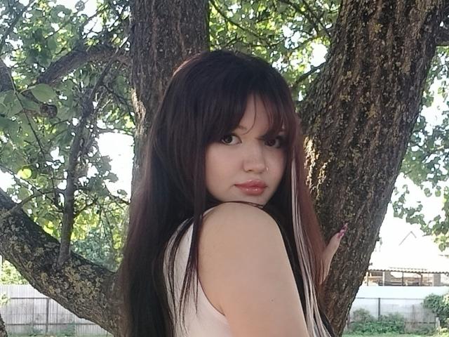 MistyEllstrom with long dark brown hair, looking back, standing by a tree, wearing a white sleeveless top, showing her ass slightly, with a nose ring and pink lips.