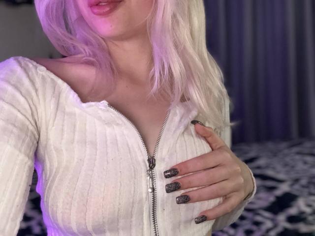 FallenAngelia with white ribbed zip-up top, hand on her tits, long blonde hair, purple-lit face, pink lips, glittery nails, soft lighting, cozy bedroom setting.