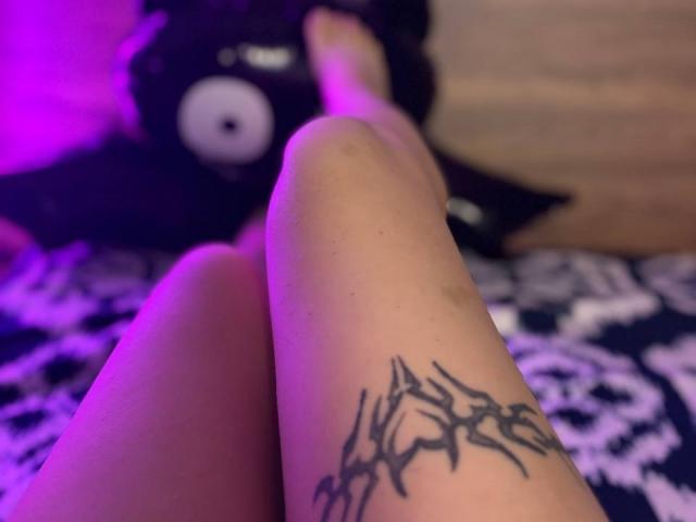 FallenAngelia's leg with a black spider tattoo, crossed at the knee, under purple lighting. A black plushie with a white eye is partially visible in the blurry background.