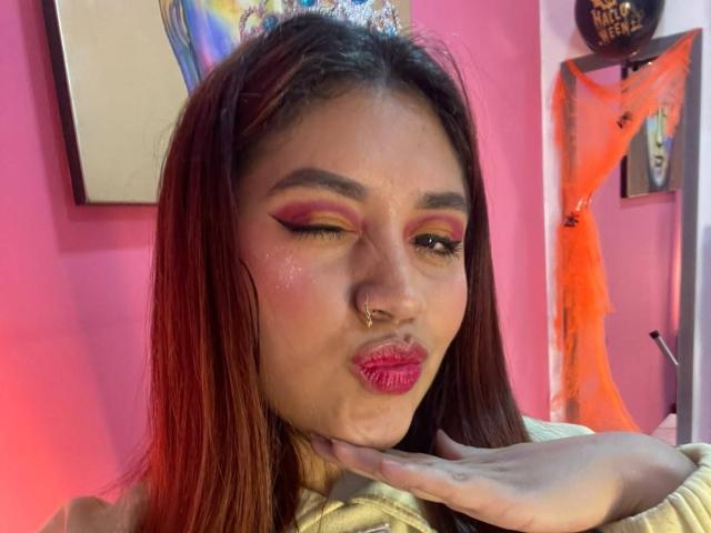 ValmontAlessia with red-brown hair, pink eyeshadow, gold lipstick, nose ring, and a hand touching her chin, wearing a golden top, pouty lips, pink walls in background.