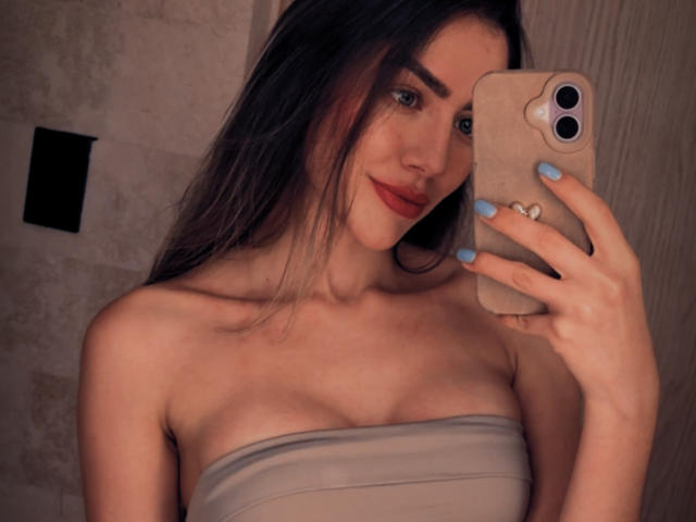 Een vrouw met lang donker haar, blauwe nagels, rode lipstick, een top zonder bandjes, die een telefoon vasthoudt, lachend, met kleine borsten, in een spiegelzelfportret, in een zwak verlichte kamer..